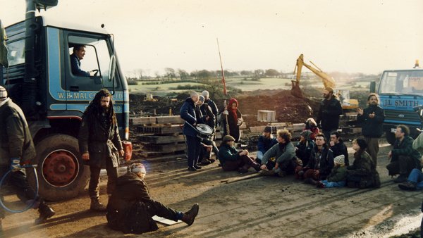 Photograph of a group of people sitting on a dirt road to block a truck.